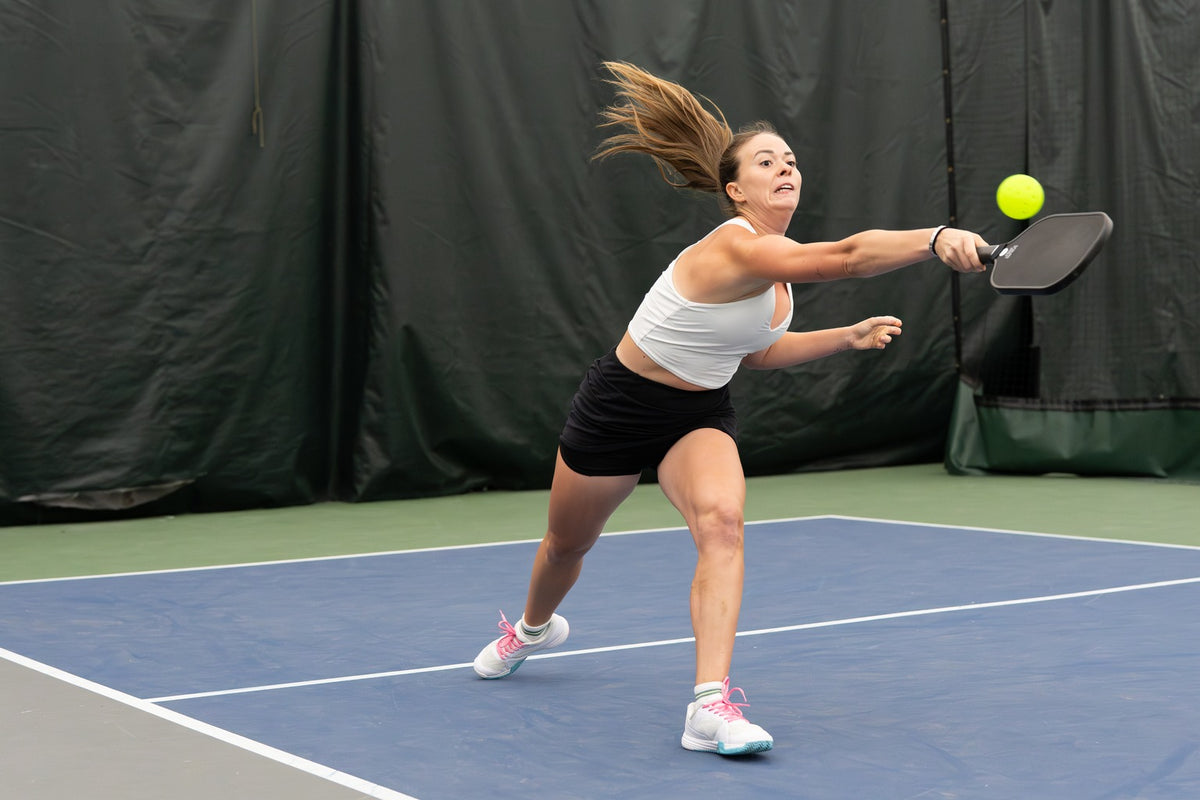 A pickleball player in a white top and black skort lunges wide on a blue indoor court, reaching her paddle toward the ball in the transition zone