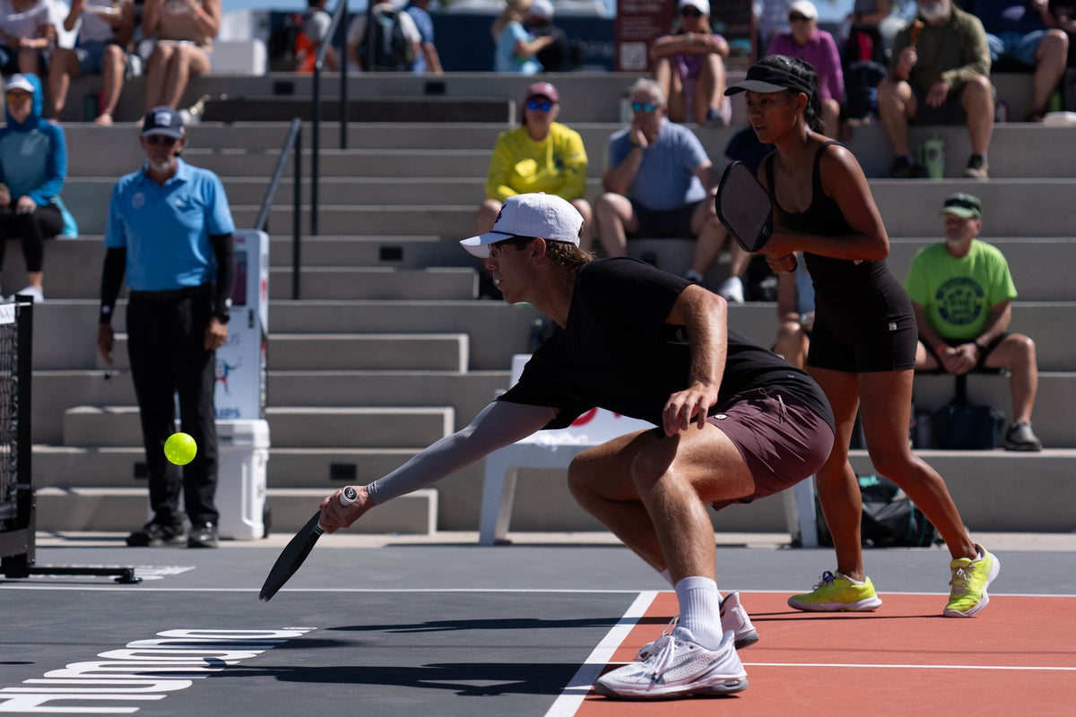 Player stretches low for a forehand at the kitchen line as his partner waits ready behind him, with a tournament crowd in the stands