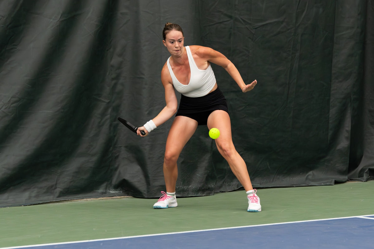Player waits behind the baseline in a low ready stance, paddle back and eyes on the incoming ball on an indoor court