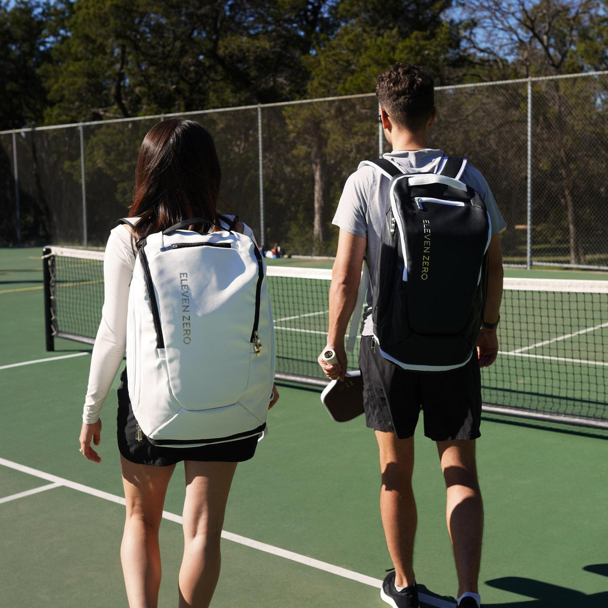 Two pickleball players walking onto an outdoor court carrying Eleven Zero backpacks and a paddle