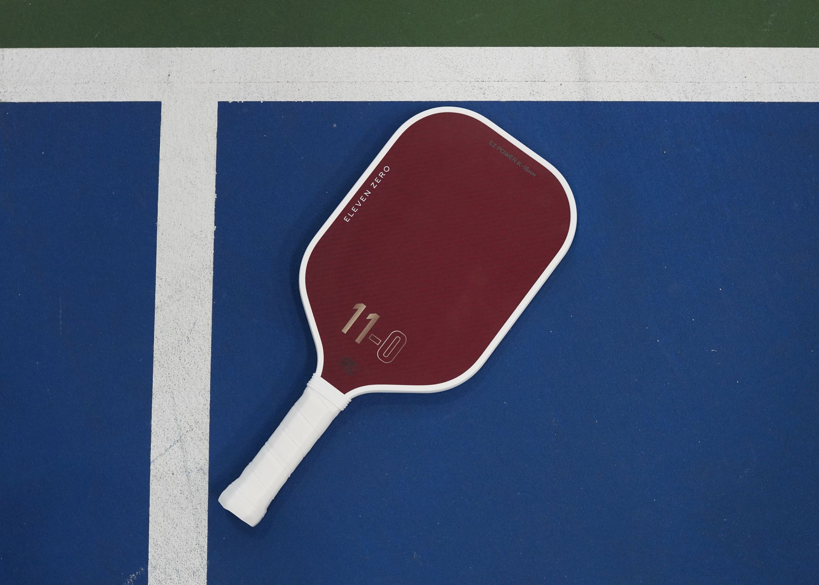 Red Eleven Zero kevlar pickleball paddle resting on a blue court with white boundary line.