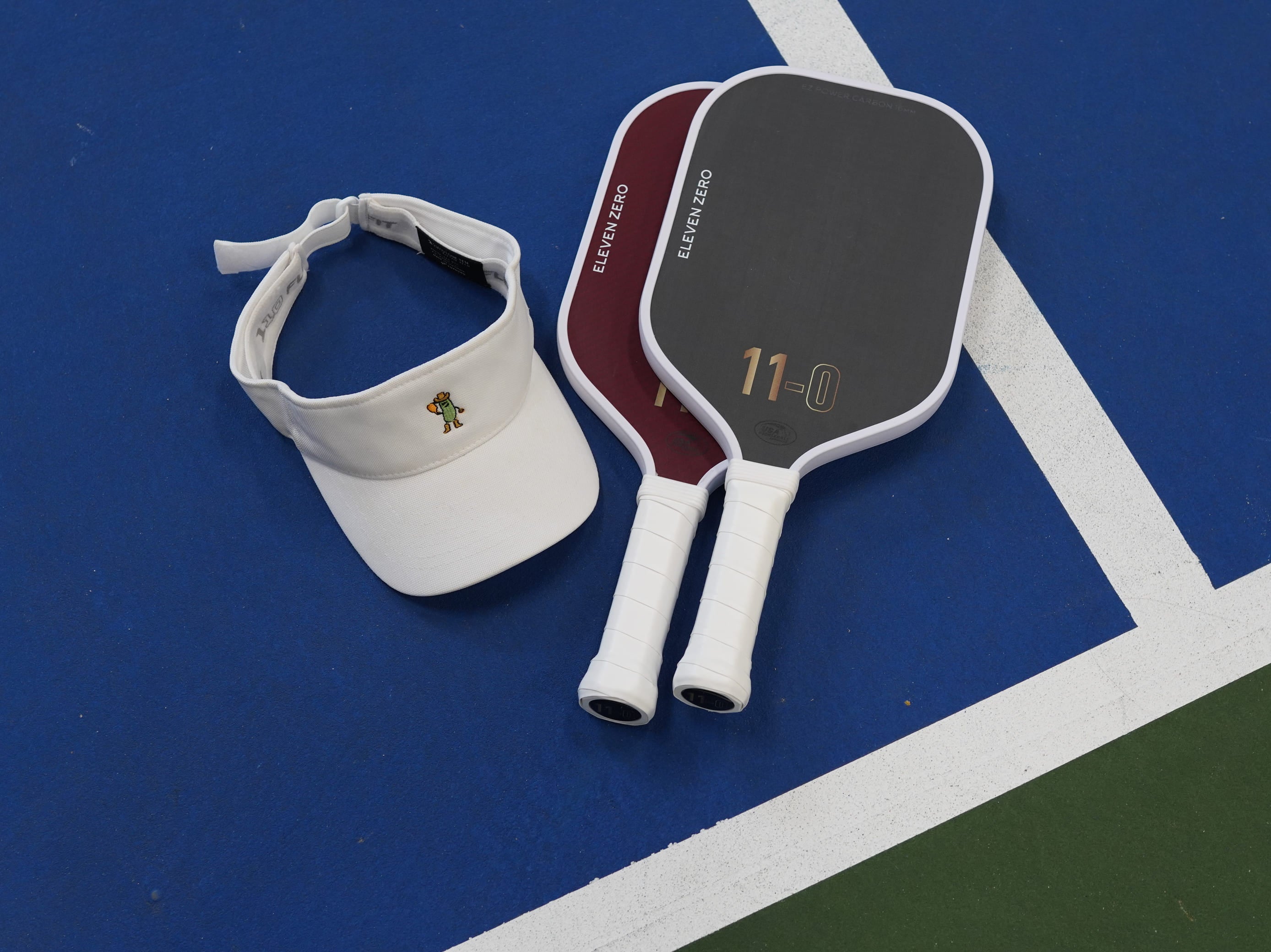 Eleven Zero carbon and kevlar pickleball paddles resting on a blue pickleball court alongside a white embroidered visor.