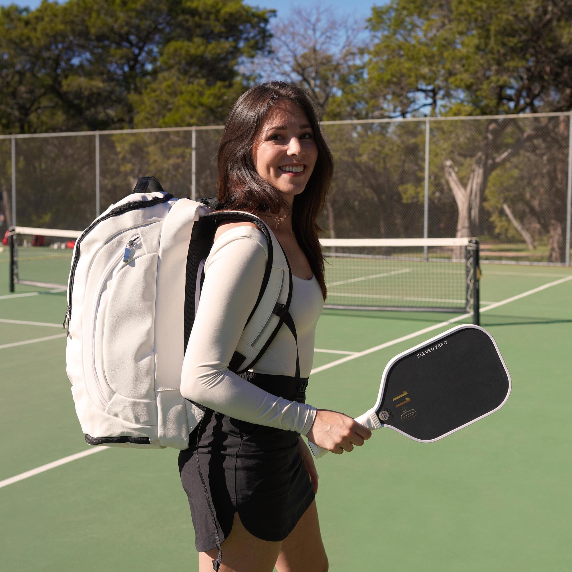 Woman on a pickleball court wearing a white Eleven Zero backpack, holding a black-and-white paddle, smiling towards the camera.