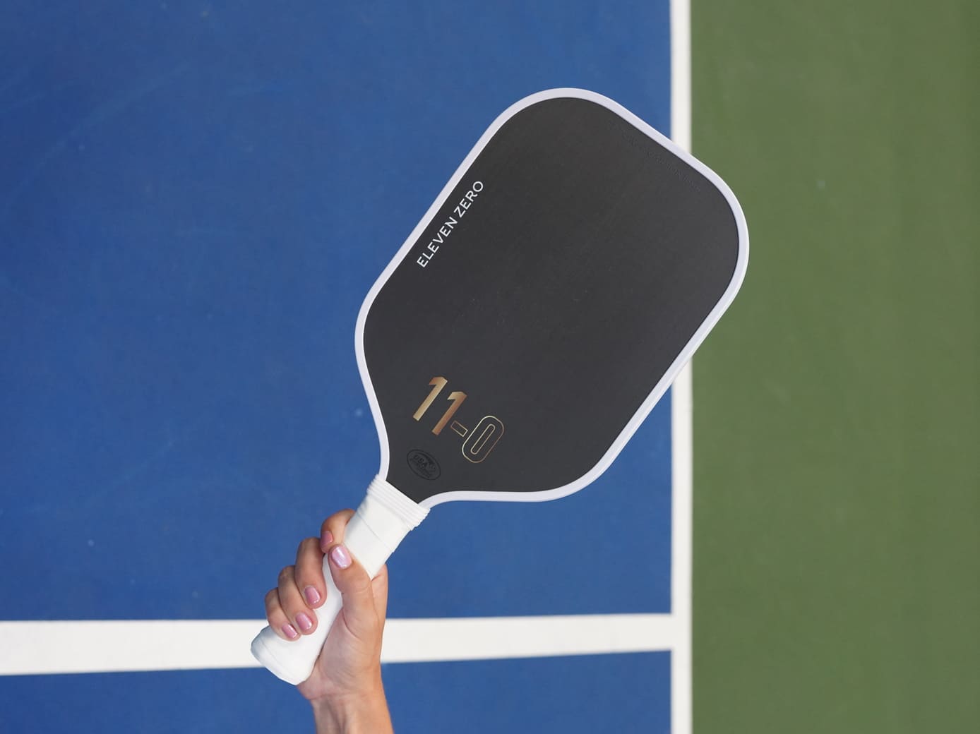 Lifestyle image of a hand holding the Eleven Zero Carbon paddle on a pickleball court, with green and blue court background