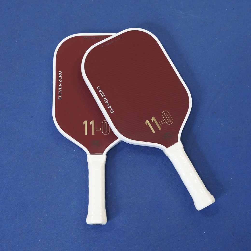 Two maroon Eleven Zero K-16 paddles on a blue court, showing front faces and handles