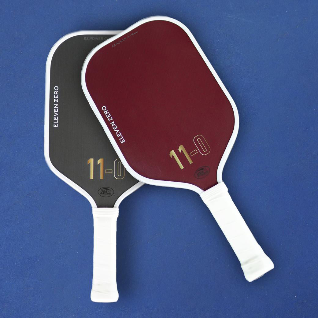 Eleven Zero Sports pickleball paddles in black carbon and Kevlar resting on a blue pickleball court surface.
