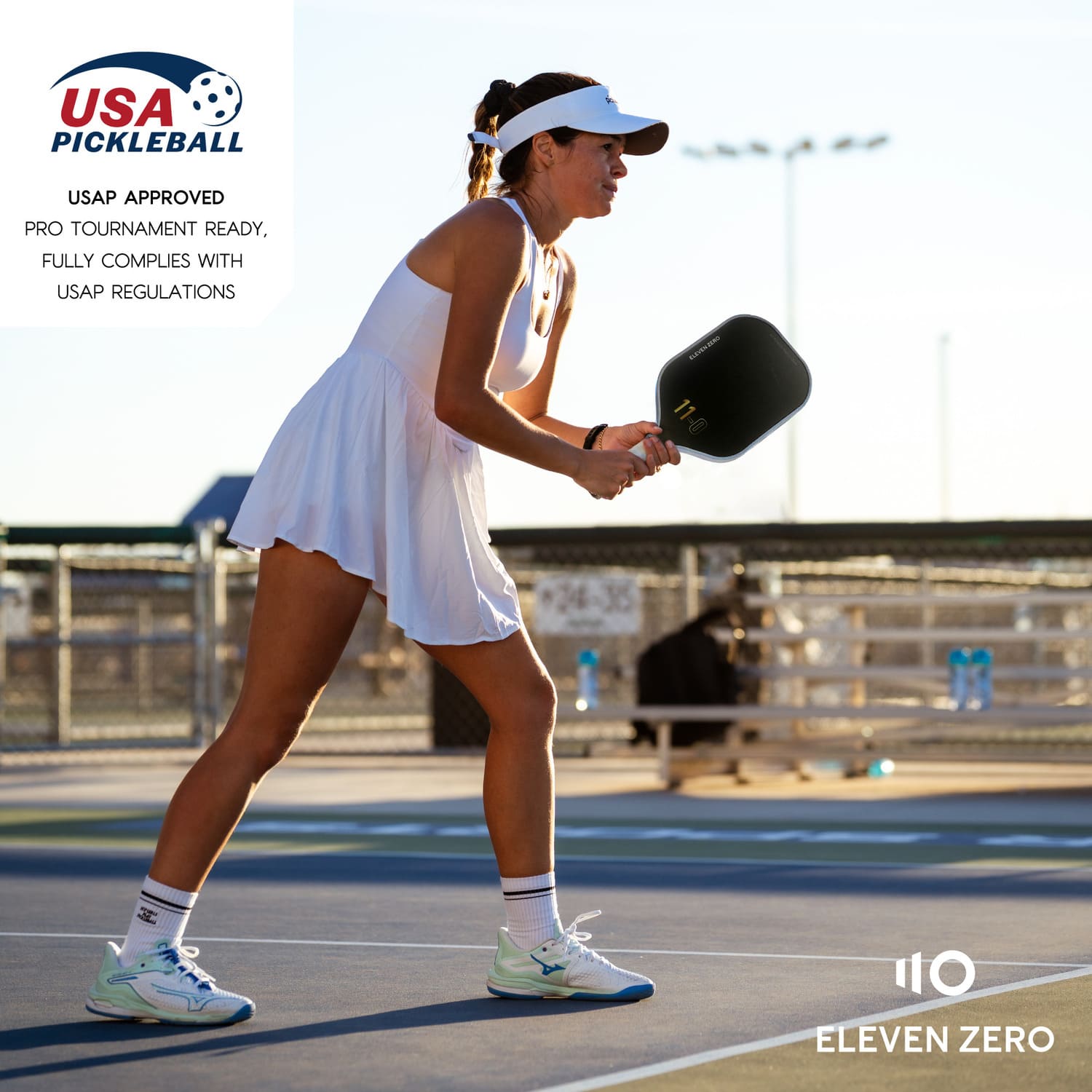A female pickleball player dressed in white athletic wear holds an Eleven Zero pickleball paddle, ready to play on a sunny outdoor court. Text indicates USA Pickleball approval, suitable for pro tournaments.