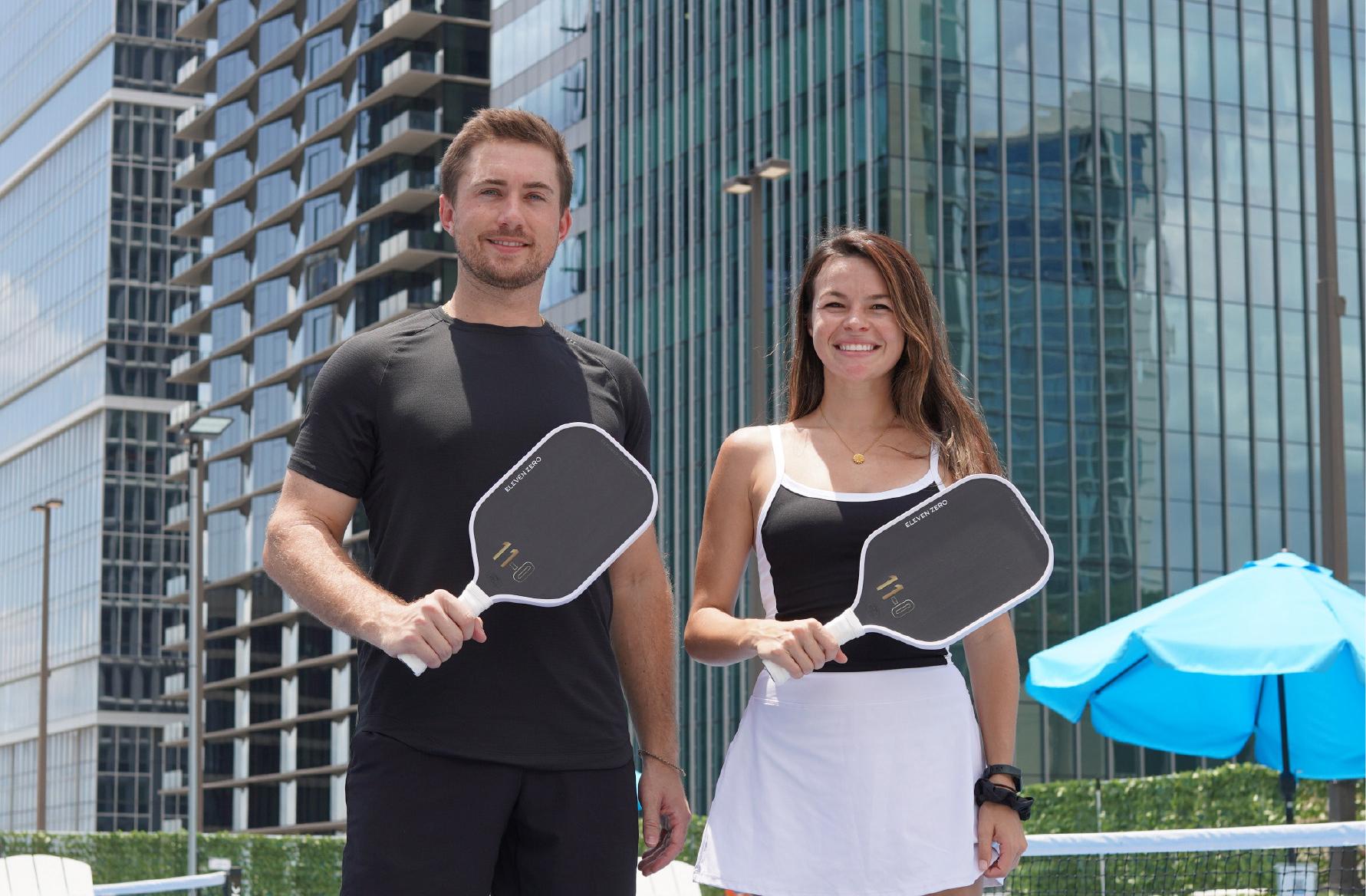 Two smiling pickleball players holding black Eleven Zero Carbon paddles, standing confidently with contemporary buildings in the background.