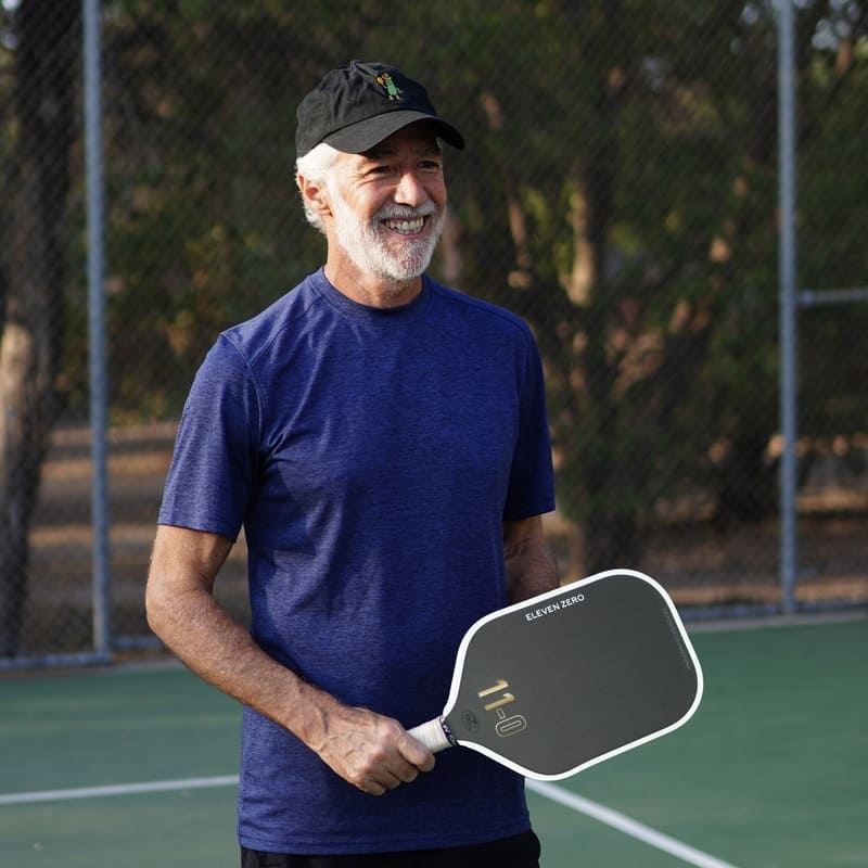 Older man smiling and holding an Eleven Zero paddle on a court, wearing a blue shirt and black hat.