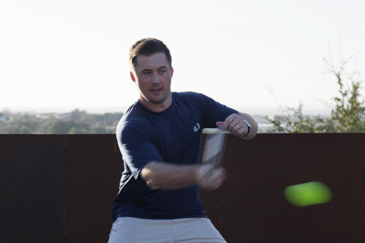 A pickleball player in a navy Eleven Zero shirt hits a forehand on an outdoor rooftop court