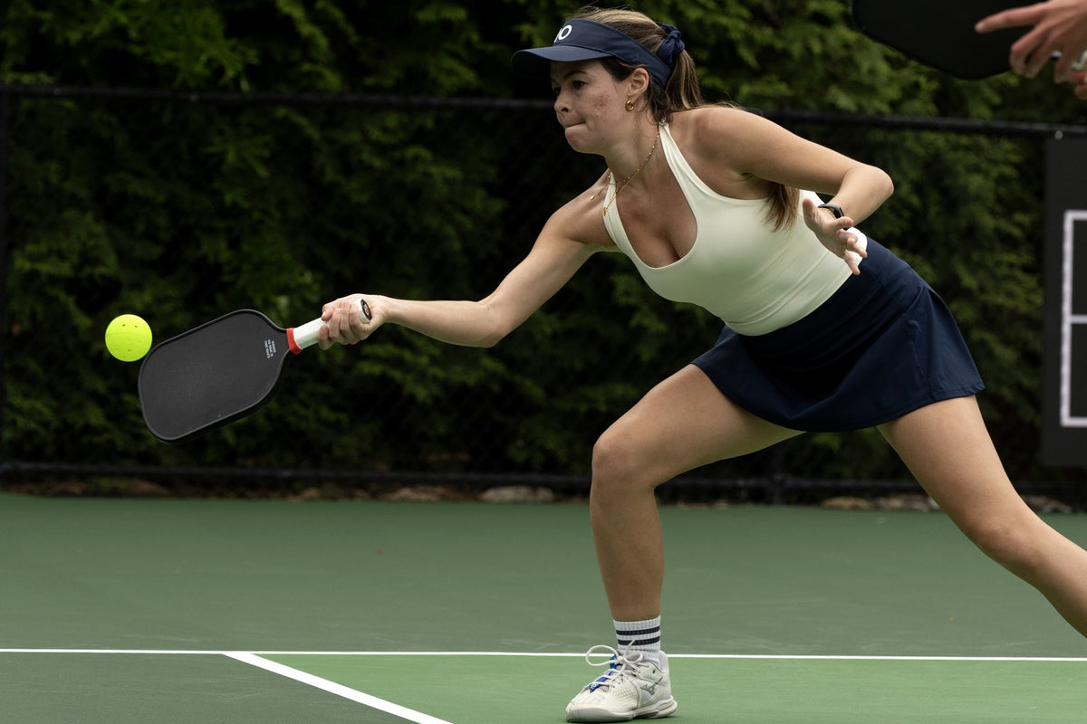 Player in an Eleven Zero visor lunges into a forehand drive with an Eleven Zero paddle, eyes on the ball, on an outdoor court