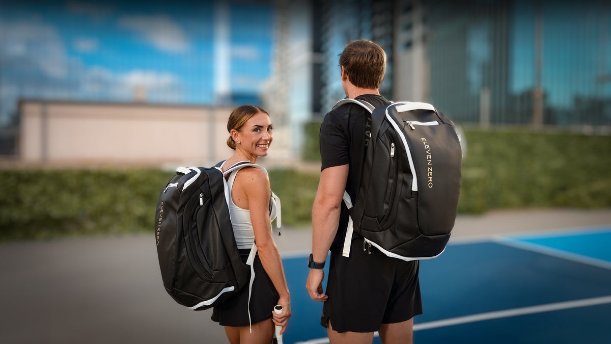 Two pickleball players carrying Eleven Zero backpacks onto a blue court with city towers behind them