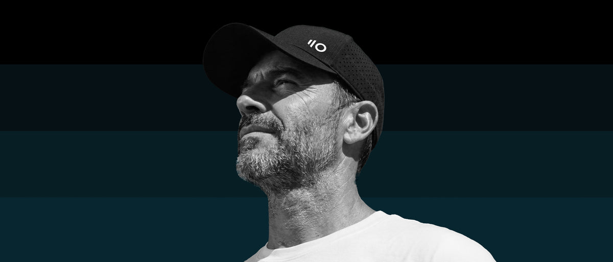 A man in profile wearing a black pickleball cap with a white 'eleven zero' logo and a white t-shirt, set against a dark teal background.