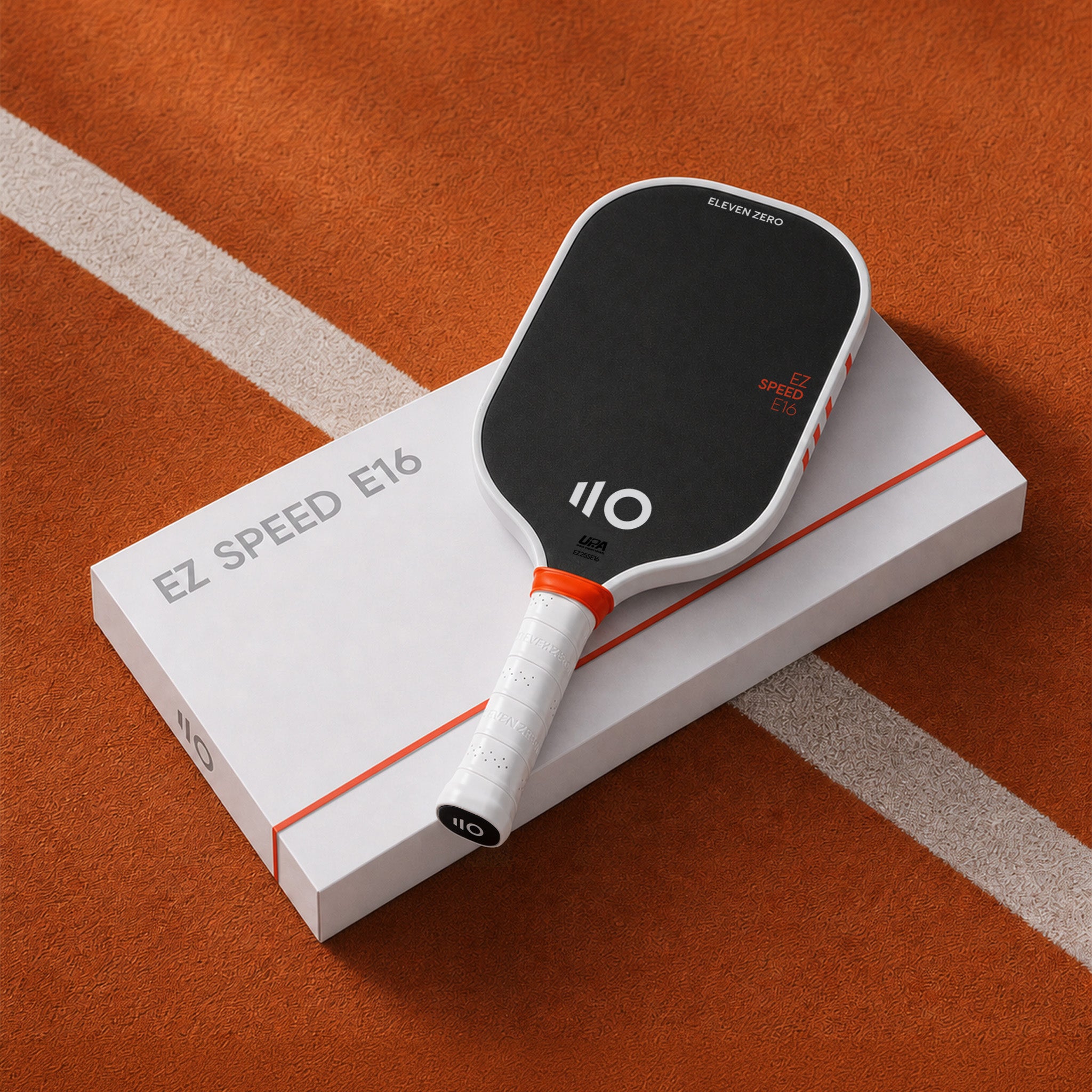 A pickleball paddle resting on top of its white retail box, photographed on an orange pickleball court surface with a white line. The paddle is black and white with orange accents, and the box is labeled EZ SPEED E16.