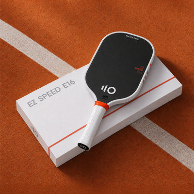A pickleball paddle resting on top of its white retail box, photographed on an orange pickleball court surface with a white line. The paddle is black and white with orange accents, and the box is labeled EZ SPEED E16.