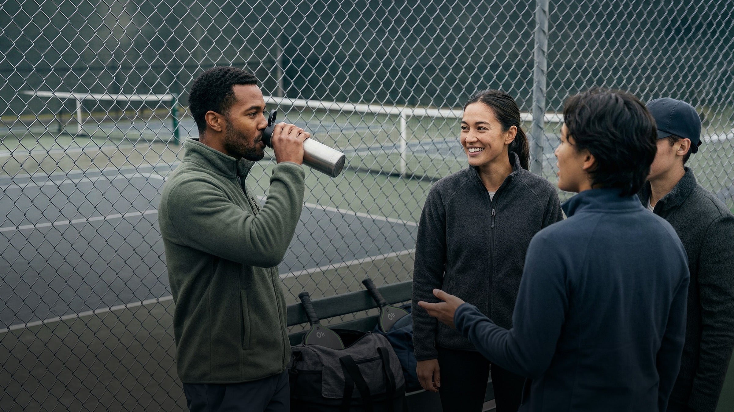 A group of pickleball players chatting after the game, with a pickleball court in the background and a pickleball bag and paddles on a bench.