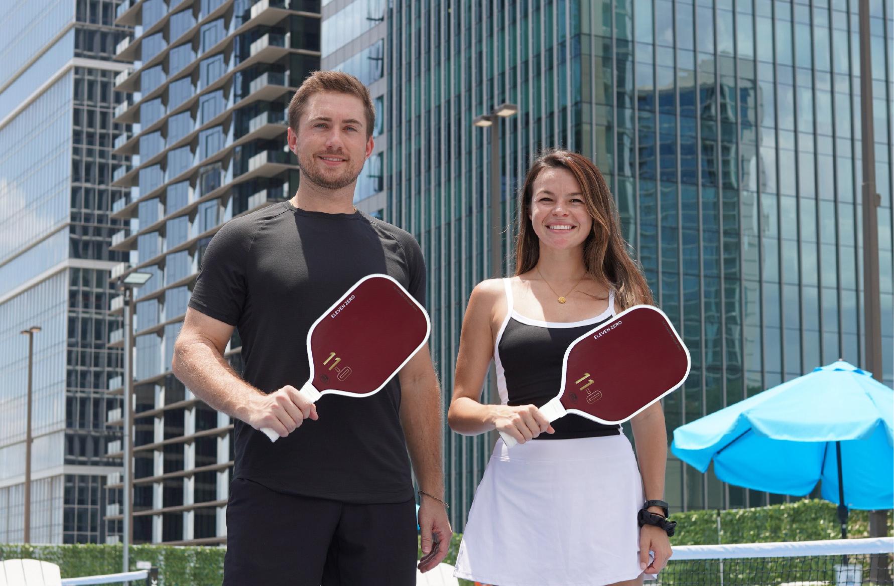 Two smiling pickleball players holding maroon Eleven Zero Kevlar paddles, standing confidently in front of modern urban buildings.
