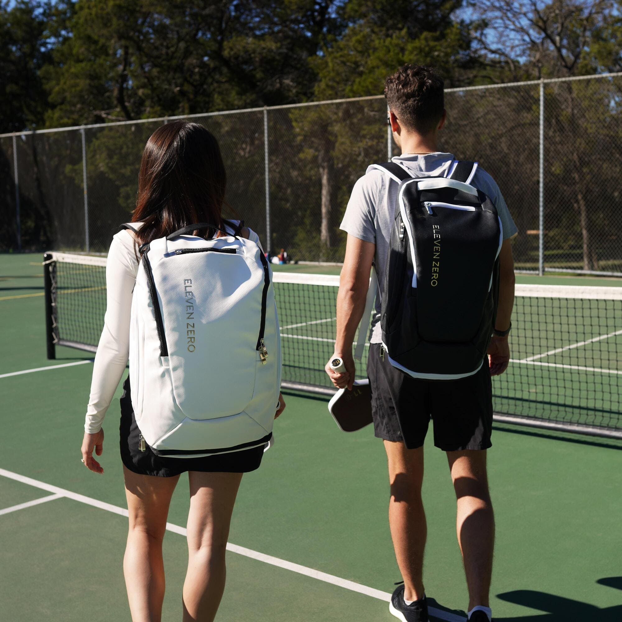 Man and woman walking on a pickleball court, wearing black and white Eleven Zero backpacks, each holding a paddle, facing the net.